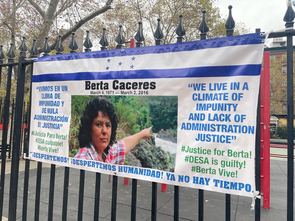
Pegados a las rejas de un parque infantil, la bandera hondureña y un cartel con la imagen de la activista Berta Cáceres, asesinada en 2016, estaban entre los indicadores que visibilizaron la resiliencia y resistencia de lxs protestantes. Foto: Dashiell Allen.
